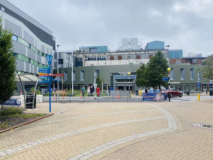 Modern hospital entrance with people walking and signs for Emergency Dept, main entrance, and ambulance bay under cloudy sky.